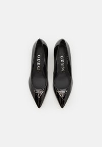 Scarpe con tacco alto in vernice nera, con punta appuntita, caratterizzate da un accento metallico triangolare con il marchio "GUESS" sul davanti.