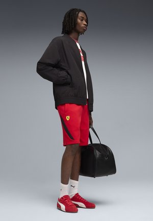 Giovane con giacca nera, pantaloncini rossi Ferrari, scarpe Puma rosse, calzini bianchi, che tiene una borsa da viaggio nera, in piedi davanti a uno sfondo grigio.