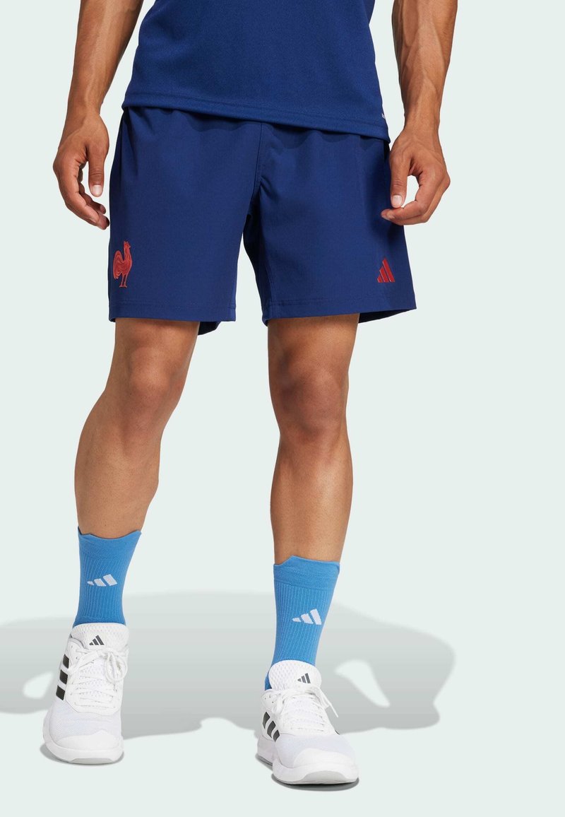 Marina atletiska shorts med röd tupp-logga och röd Adidas-logotyp. Matchas med blå strumpor och vita sneakers med svarta ränder.