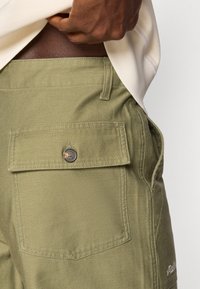 Shorts cargo verde măsliniu cu un buzunar mare la spate, dotat cu închidere cu buton. Realizați dintr-un material texturat cu detalii de cusătură vizibile.