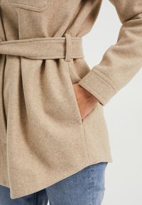 Béžová vlna-směsová bunda s opaskem v pase, bočními kapsami a uvolněným střihem. Jaklový design s jemným, strukturovaným povrchem.