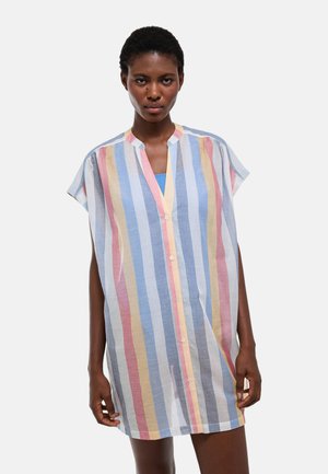 Femme portant une robe ample boutonnée avec des rayures verticales dans des tons pastel bleu, jaune, rose et blanc, debout devant un fond uni.