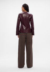 Burgundská kožená sako s priliehavým strihom a dlhými rukávmi, v kombinácii so voľnými hnedými nohavicami s vreckami na chrbte a rovnými nohavicami.