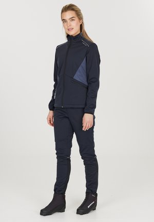 Marineblaue Softshelljacke mit hohem Kragen, dreieckigem Paneeldesign, mit Reißverschlusstaschen, kombiniert mit passenden Hosen und schwarzem Schuhwerk.