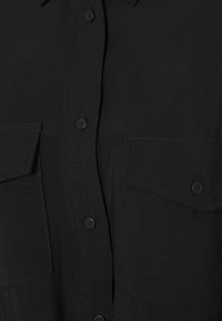 Devant d'une chemise noire avec fermeture boutonnée et deux poches poitrine, chacune sécurisée par un rabat à bouton unique.