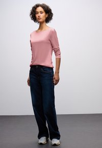 Světle růžové dlouhé tričko v kombinaci s tmavě modrými širokými denimovými kalhotami a šedými teniskami, na modelce před jednoduchým pozadím.