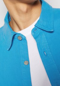 Camicia in denim blu con colletto, bottoni a pressione argentati e finitura Texturizzata. Indossata sopra una T-shirt bianca semplice.