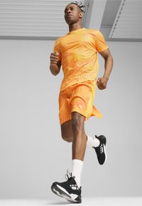 Tenue de course en orange vif avec des rayures ondulées ; comprend un haut à manches courtes et un short. Chaussures de sport noires avec des accents blancs.