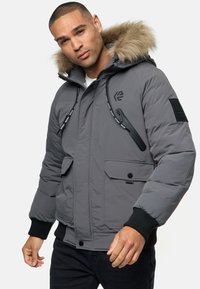 Grau gepolsterte Jacke mit einer mit Fell gefütterten Kapuze, Reißverschluss- und Druckknopftaschen, elastischen Bündchen und einem Logopatch am linken Ärmel.
