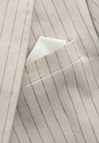 Beige Nadelstreifen-Blazer mit einem ordentlich gefalteten weißen Einstecktuch, das in die Brusttasche gesteckt ist.