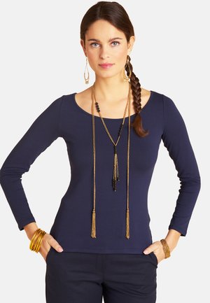 Woman with braided hair wearing a navy long-sleeve top, layered gold and black bead necklaces, and gold bracelets with hands in pockets.