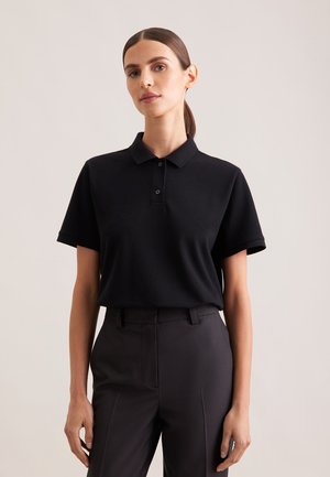 Kvinde med mørkt hår sat tilbage iført en sort kortærmet polo og sorte, skræddersyede bukser mod en ensartet lys baggrund.