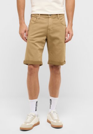 Mann trägt hochgekrempelte beige Shorts, weiße Socken mit schwarzem Text, beige-weiße Sneakers, steht vor einem schlichten hellen Hintergrund.