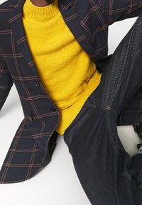 Marineblauer kariertes Blazer mit orangefarbenen Linien über einem gelben strukturierten Pullover, kombiniert mit dunklen Jeanshosen mit dezenten Stickdetails.