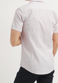 Man in een lichtgekleurde, kortafgeknotte shirt met kleine geometrische vormen, gecombineerd met donkere broek, gezien van achteren.