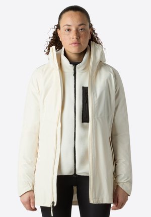 Persona con capelli ricci che indossa un cappotto invernale con cappuccio color crema sopra una giacca in pile bianca con dettagli della cerniera neri.