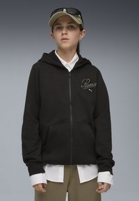 Čierna mikina na zips s predným vreckom, ribovanými manžetami a vyšitým logom "Puma". Nosená cez bielu košeľu s golierom a khaki nohavicami.