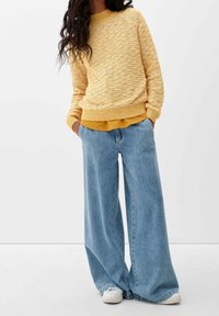 Femme portant un pull jaune texturé sur une chemise moutarde, un jean bleu clair à jambes larges et des baskets blanches, les mains dans les poches.