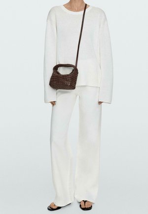 Person trägt einen übergroßen weißen Pullover und weiße Hose mit schwarzen Sandalen und trägt eine kleine dunkelbraune geflochtene Umhängetasche.