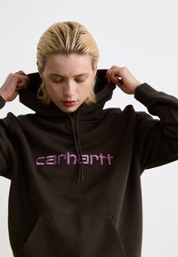 Bruine hoodie met een voorzak en een verloop van roze en paarse logotekst op de borst. Zacht, gestructureerd materiaal.