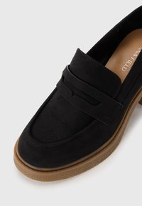 Mocassin en cuir suédé noir avec un design contour, doté d'une sangle décorative et d'une semelle en caoutchouc texturée de couleur brun clair pour une meilleure adhérence.