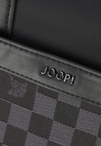 Sort ternet mønster pung med glatte læderdetaljer og sølvhardware, der har mærkenavnet "JOOP!" præget på forsiden.