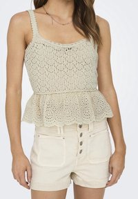 Top de crochet en beige claro con dobladillo en forma de ondas, cuerpo ajustado y tirantes finos, combinado con pantalones cortos de cintura alta en color crema.