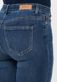 Pogled od blizu na osebo, ki nosi temno modre kavbojke z usnjenim labelom "ONLY" na pasu.