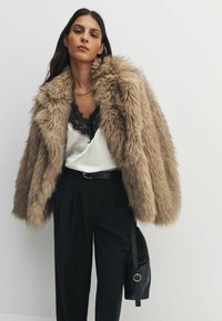 Faux ādas mēteli gaiši brūnā krāsā, ar mīkstu tekstūru un pārmērīgi lielu dizainu. Kombinēts ar melnu mežģīņu apmalē tērpu un piegriezuma melniem biksēm.