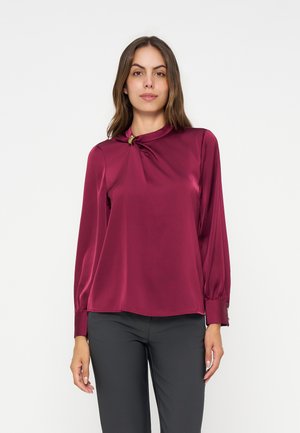 Femme portant un chemisier bordeaux brillant à manches longues avec un col froncé et un pantalon noir sur un fond clair uni.