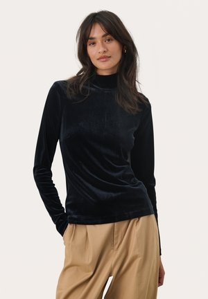 Svart sammet långärmad topp med hög halsringning, slät textur och en figurnära design. Kombinerad med lösa beigefärgade byxor.