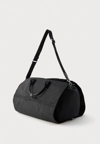 Mustasta kankaisesta kassi-matkalaukusta, jossa on kaksi lyhyttä kahvaa ja säädettävä pitkä olkahihna, valkoista taustaa vasten.