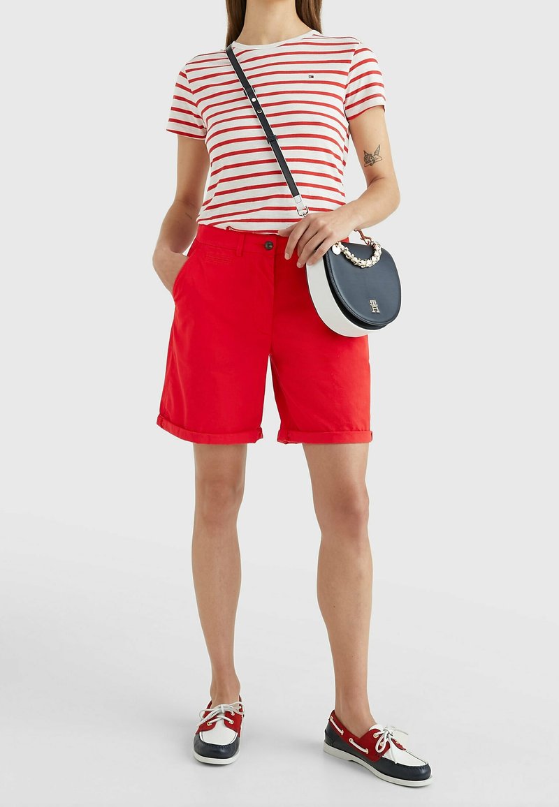 Femme portant un short rouge, un t-shirt rayé blanc et rouge, des mocassins bateau tricolores et un sac bandoulière noir et blanc avec une chaîne dorée.
