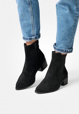 Zwarte fluwelen enkellaarsjes met een spitse neus, een zijrits en een lage blokhak, gedragen met opgerolde lichtblauwe jeans.