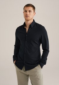 Temno modra srajca s gumbi iz gladke tkanine, z ostrim ovratnikom in dolgimi rokavi. Majhen logotip na prsih. Hlače nevtralne barve.
