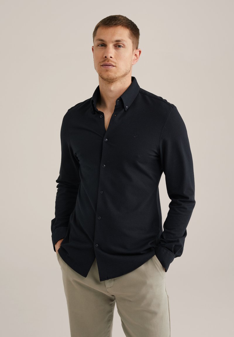 Temno modra srajca s gumbi iz gladke tkanine, z ostrim ovratnikom in dolgimi rokavi. Majhen logotip na prsih. Hlače nevtralne barve.