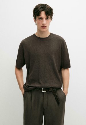 Jonge man met kort bruin haar, gekleed in een donkerbruine kortärmelige gebreide trui en bijpassende broek met een gevlochten riem, handen in de zakken.