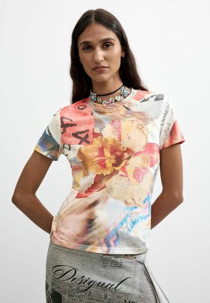 Femme aux longs cheveux foncés portant une chemise colorée à motifs floraux et texte, ainsi qu'un collier ras-de-cou floral, debout sur un fond blanc.