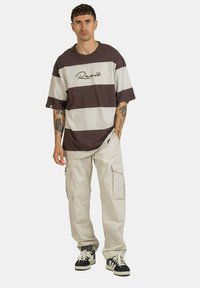 Bruin en beige gestreepte oversized t-shirt met bedrukte logo, gecombineerd met crèmekleurige cargobroek en zwart-witte sneakers.