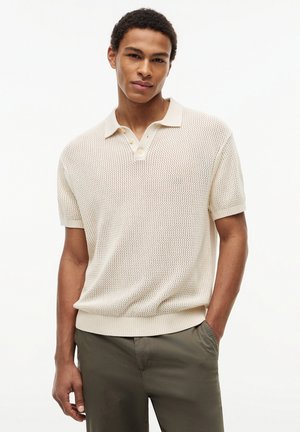 Junger Mann trägt beige kurzärmliges, gestricktes Poloshirt und olivgrüne Hose, steht mit einer Hand in der Tasche vor weißem Hintergrund.