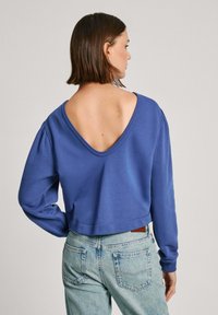 Zils džemperis ar dziļu V formas aizmuguri, garām piedurknēm, ribotām aprocēm un saīsinātu apakšu, kombinēts ar gaišiem džinsu biksēm. Gluda kokvilnas auduma.