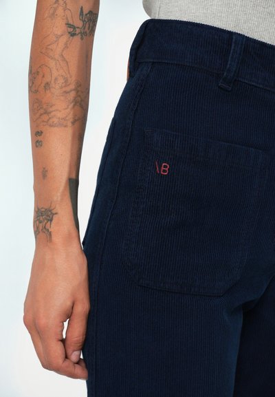 Pantaloni din velur navy cu o suprafață texturată, având un buzunar pe spate și un mic logo roșu brodat pe buzunar.