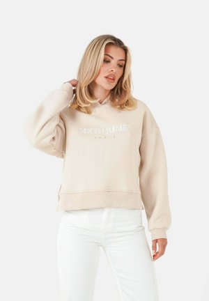 Beige hoodie with a loose fit, featuring a white embossed logo "SIXTHJUNE" on the front. Soft fabric and ribbed cuffs. Worn with white jeans.