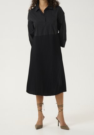 Mujer con un vestido negro de manga larga con falda plisada y tacones altos de cordones en verde oliva, de pie contra un fondo liso.