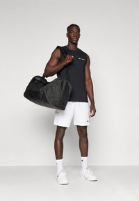 Black duffel bag with rounded edges and dual handles, made of durable fabric, held by a person in a sleeveless top and shorts.