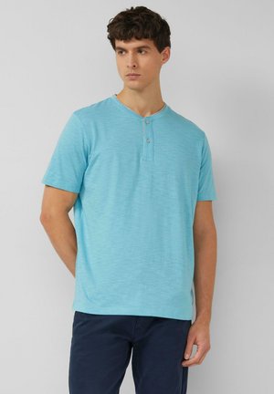 Młody mężczyzna w jasnoniebieskiej koszulce henley z krótkim rękawem i ciemnoniebieskich spodniach, stojący na tle jednolitego jasnoszarego tła.