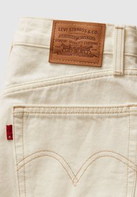 Τζιν από βαμβακερό τζιν σε κρεμ χρώμα με δερμάτινο πίσω σημείο, με πορτοκαλί ραφές και κόκκινη ταμπέλα, τονισμένα από καμπύλα ραφές στις πίσω τσέπες.