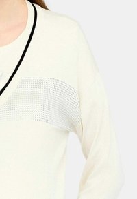 Maglione bianco con scollatura a V e un accento argentato testurizzato sul petto con dettagli a maglia sottili. Bordo nero lungo la scollatura.