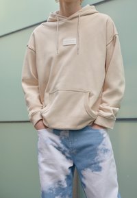 Beige huppari, jossa edessä tasku, valkoinen logopatch ja hopeanväriset kiristysnauhat, yhdistetty sinisiin ja valkoisiin pilvikuviollisiin farkkuihin.