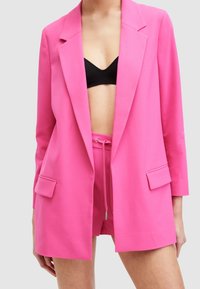 Rosa blazer med långa ärmar, snedskurna slag och två framfickor, matchad med shorts och en svart topp. Slät tygtextur.